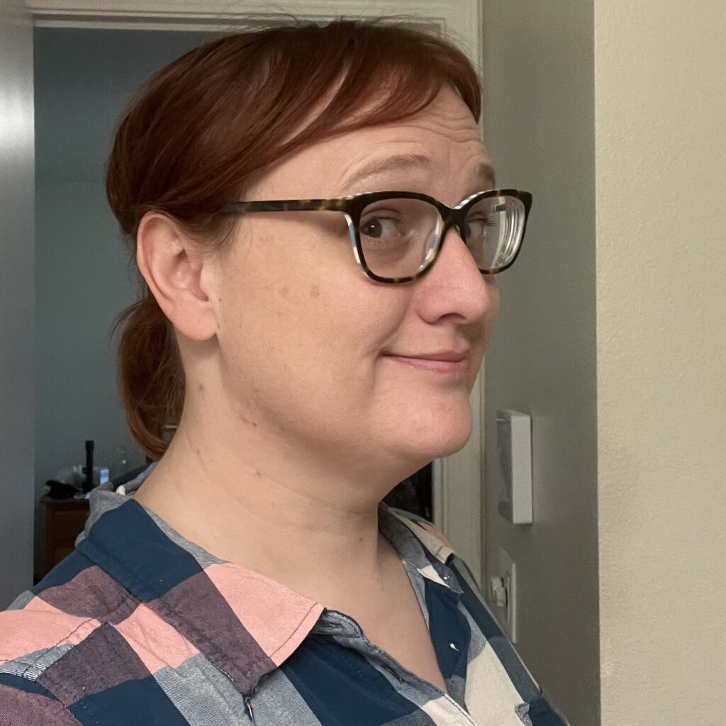 A portrait photo of the author looking at the camera. She is wearing a plaid shirt in trans pride colors, and has her eyebrows raised slightly. Her hair is up ina. ponytail.