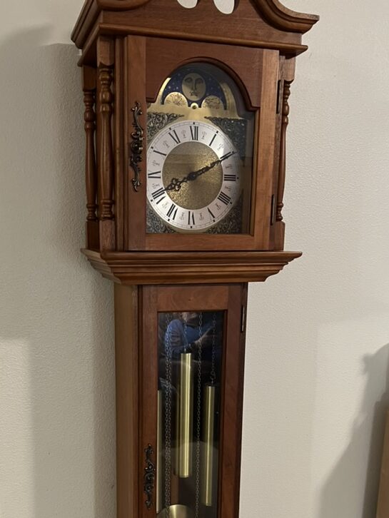 A Tall Grandfather Clock made by Franz Hermle of West Germany. The Case is tall and made of wood, while three heavy weights and a pendulum sit in the lower compartment. A dial and moon dial sit in the upper compartment.